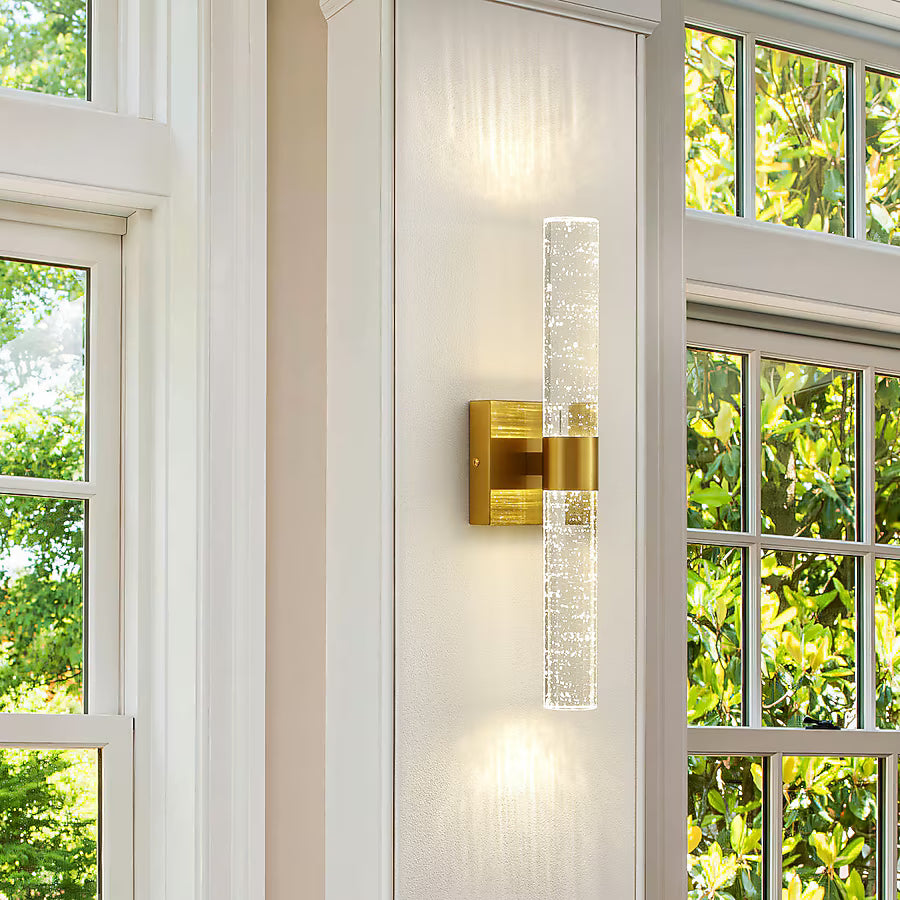 Modern wall sconce with gold finish and clear glass on a white wall with windows in the background.