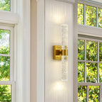 Modern wall sconce with gold finish and clear glass on a white wall with windows in the background.