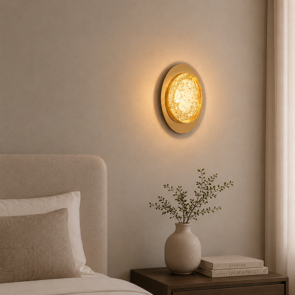Round wall light fixture on a beige wall above a nightstand with a vase and books.